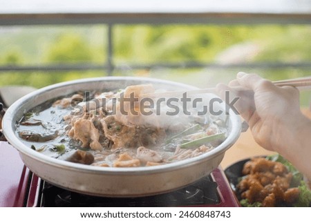 Slice of pork was picking up by the chopstick in the woman's hand above the curved brass pan around with diffused smoke. Royalty-Free Stock Photo #2460848743