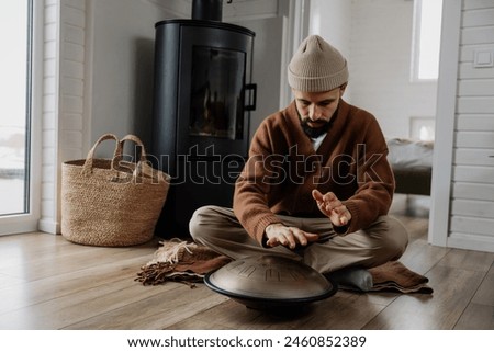 young bearded man with a melodious musical instrument that emits cosmic sounds: glucophone or handpan, stylish photo of playing tongdrum in the interior of a white wooden house
​