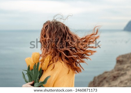 Portrait of a happy woman with hair flying in the wind against the backdrop of mountains and sea. Holding a bouquet of yellow tulips in her hands, wearing a yellow sweater