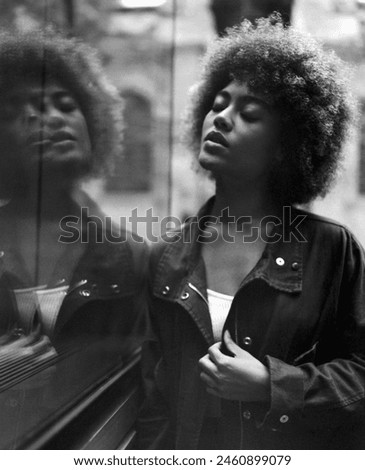 Shaken monochrome image of young woman leaning on glass wall with her eyes closed in a relaxed way. Analogue film photography. She is wearing a jacket. Grainy image.