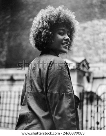 Grainy monochrome image of smiling curly young woman looking at camera. It is a back view and she is walking. Urban vibes, analog film camera.