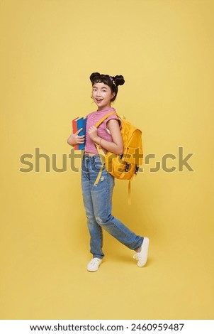 Portrait of happy smiling young Asian student elementary school girl with notebook and backpack ready to school isolated on yellow background.