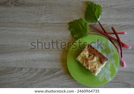 Rectangular piece of spring rhubarb pie on a plate. A plate with dessert on the table. Nearby are rhubarb stems and leaves. Gastronomy