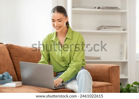 A woman is seated on a couch, engaged with a laptop computer placed on her lap. She is focused on the screen, typing and scrolling