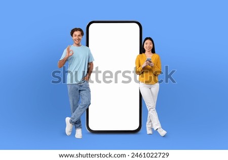 Multiracial lovers standing next to a massive phone prop isolated on blue studio background. They are looking at the phone and appear engaged in conversation or observation. Royalty-Free Stock Photo #2461022729