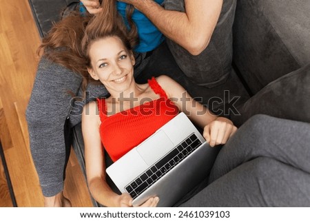 Smiling woman lying on a sofa in a red top, holding a laptop, with man in the background. A relaxed, homey setting reflecting technology use and couple goals.