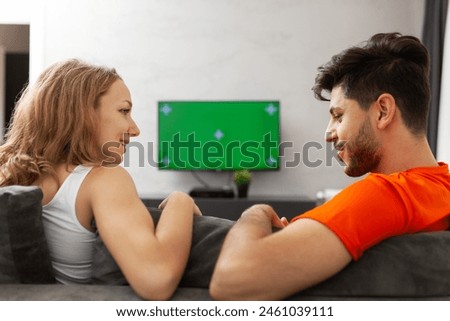A young woman and man sit on a gray couch, smiling at each other, enjoying a relaxed and joyful moment with a green screen television in the background.