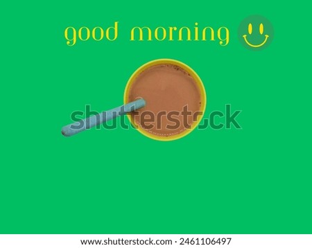 Hot chocolate in a yellow cup with a blue spoon in it can be seen from above in the picture. with "Good morning" written in white on a green background.
