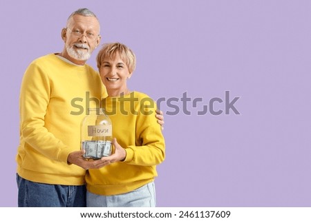 Mature couple holding jar with money for house on lilac background