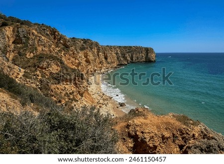 Rocky cliffs along a coastline with a secluded sandy beach. The clear turquoise waters gently lap against the shore under a bright, clear sky.Lush vegetation dots the cliffs, enhancing the scenic view