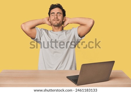 Hispanic young man working on laptop feeling confident, with hands behind the head.
