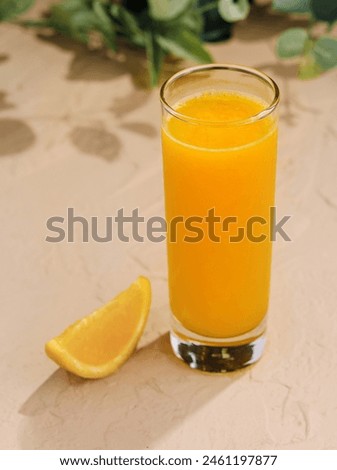 Vibrant glass of orange juice beside a fresh citrus slice on a sandy texture background with foliage shadows