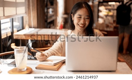 A young positive, happy Asian female college student studying online and doing homework at a coffee shop in the city, taking notes in her notebook, participating online lecture, working remotely. Royalty-Free Stock Photo #2461214937