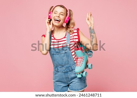 Young happy woman wear red t-shirt denim overalls casual clothes hold blue rollers listen to music in headphones sing isolated on plain pastel pink background. Summer sport lifestyle leisure concept
