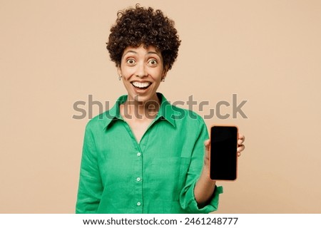 Young surprised woman of African American ethnicity wear green shirt casual clothes hold use mobile cell phone with blank screen workspace area isolated on plain beige background. Lifestyle concept