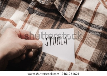 White card with a handwritten inscription "Studia", held in the hand against the background of a brown plaid shirt (selective focus), translation: study
