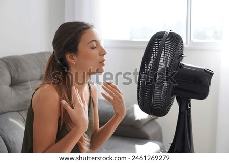 Summer heat. Young Hispanic woman cooling down by ventilator at home, feeling unwell with high temperature during hot weather. Latin girl in front of electric fan during extreme heatwave. Royalty-Free Stock Photo #2461269279