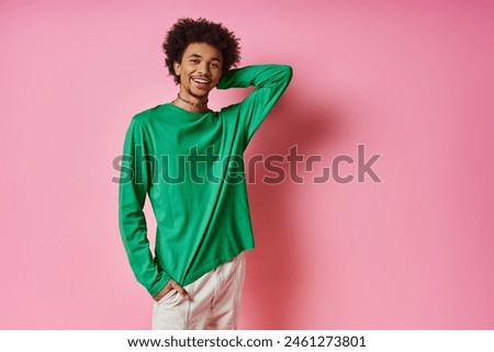 A cheerful, curly-haired African American man in a green shirt strikes a pose for a picture on a pink background.