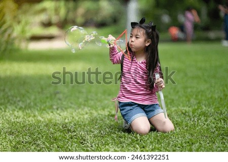 A young Asian girl in a striped shirt plays with a giant bubble wand outdoors, creating large, colorful bubbles.

