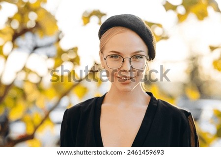 The face of a beautiful girl with light daytime makeup, transparent glasses on her eyes and a black beret, wearing a black blouse against the background of a parking and autumn leaves on a tree