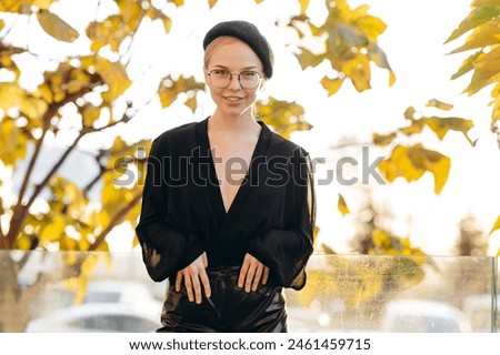 Portrait of a sophisticated girl with light daytime make-up and bun hair, with clear glasses on her eyes and a black beret on her head, holding her hand on her head, wearing a black blouse