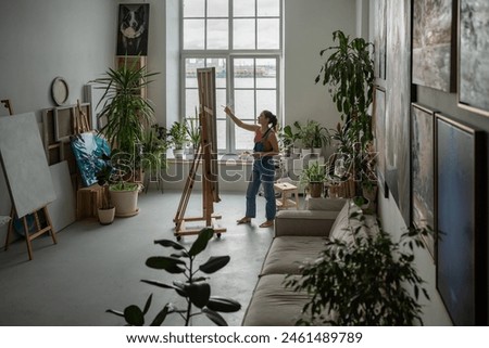 Creative female artist paints with oil on canvas, standing near easel with brush. Focused woman creating picture, making artwork at home. Inspired girl at workshop doing art with acrylic paints.