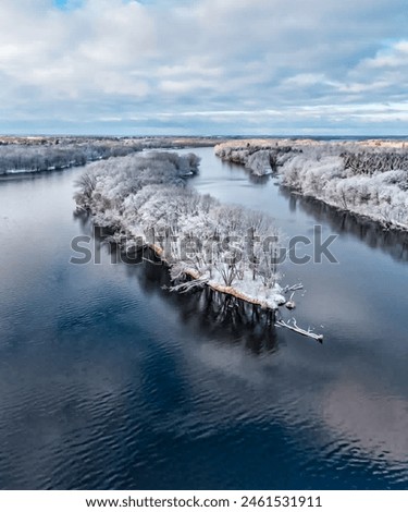 Serene winter landscape with a tranquil river flowing through a frosty forest, reflecting the cloudy sky. Snow-covered trees and icy waterscape create a beautiful, cold, and serene outdoor scenery 