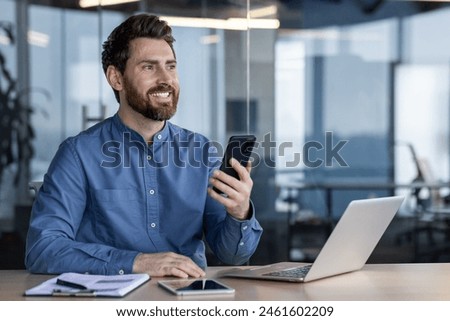 A young man works in the office, sits at a desk and uses a mobile phone, smiling and looking ahead. Close-up photo.