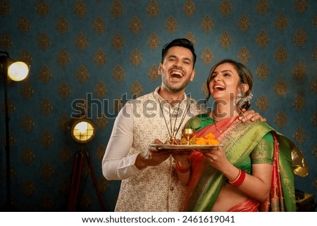 Picture of a sweet pair celebrating Diwali while dressed in traditional Indian attire, holding a puja dish in their hands and smiling
