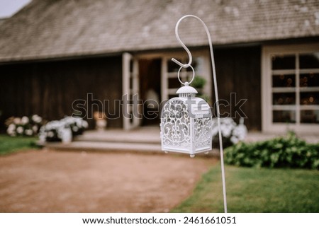 Valmiera, Latvia - August 25, 2023 - Decorative white lantern hanging on a hook in a rustic outdoor setting, with a blurry background of a wooden building.