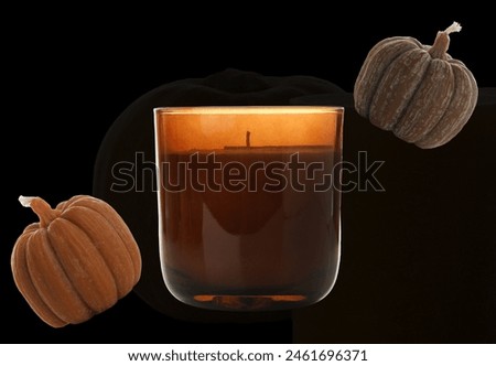 a dark amber candle in a glass holder, accompanied by two small, textured pumpkins with a dusty appearance. The dark background enhances the warm, autumnal ambiance.