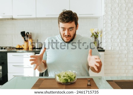 A young man sits in the kitchen in front of a glass bowl of salad with mixed feelings. He looks at the salad with bulging eyes. Greens again! Royalty-Free Stock Photo #2461715735