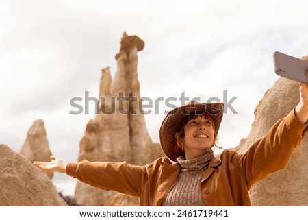 A woman in a brown jacket and hat is taking a picture of a mountain. She is smiling and she is enjoying the scenery