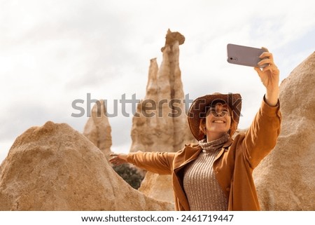 A woman in a brown jacket and hat is taking a picture of a mountain. She is smiling and she is enjoying the scenery
