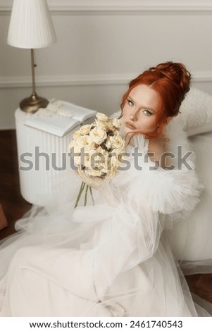 A beautiful lady with fiery red hair elegantly holding a bunch of white roses in a white attire indoors