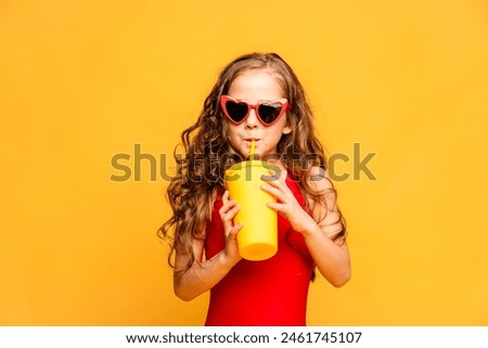 Little smiling beautiful girl in sunglasses and red swimsuit drinking lemonade through a tube on yellow background, Summer, beach concept. Copy space. high quality photo