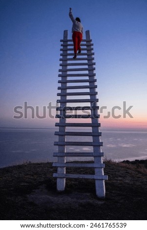 A young woman has raised her hand and is standing at the top of a wooden staircase outdoors. Stairs to the sunset sky on the background of the seascape. Success, growth and goal achievement concept.