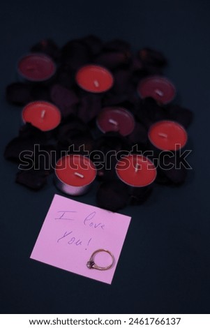 "I love you" note in a pink paper with a gold ring, dried flowers leaves and candles on a black background.
