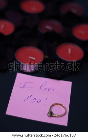 "I love you" note in a pink paper with a gold ring, dried flowers leaves and candles on a black background.