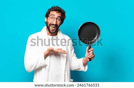 young crazy bearded man wearing a bathrobe cooking with a pan Royalty-Free Stock Photo #2461766531