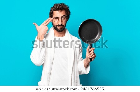 young crazy bearded man wearing a bathrobe cooking with a pan Royalty-Free Stock Photo #2461766535