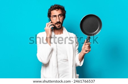 young crazy bearded man wearing a bathrobe cooking with a pan Royalty-Free Stock Photo #2461766537