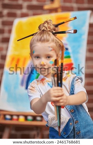Children's creativity. Portrait of a cheerful little girl, stained with colorful paints and with brushes in her hands, against an easel with a child’s drawing. Drawing and kids imagination. Education.