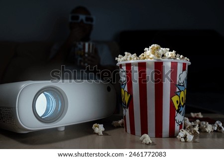 movie night at home, with a projector, a bowl of popcorn and a man in the background with three-dimensional glasses watching the movie.