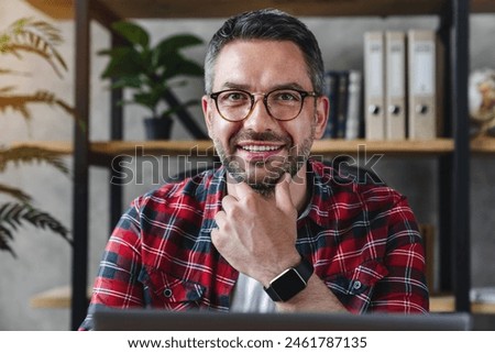 Head shot close up portrait of positive smiling caucasian freelancer businessman man in eyeglasses wearing smart watch casual outfit sitting at home office desk using laptop with look at camera
