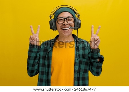 Smiling Asian man in a beanie and casual clothes throws up a peace sign while listening to music or a podcast on his headphones. Isolated on a yellow background.