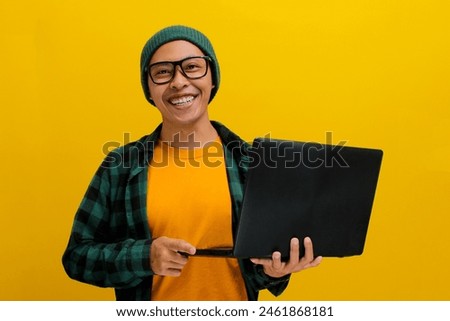 Happy Asian man, dressed in a beanie hat and casual shirt, exudes confidence and joy as he holds a laptop, smiling brightly while looking directly at the camera against a yellow background