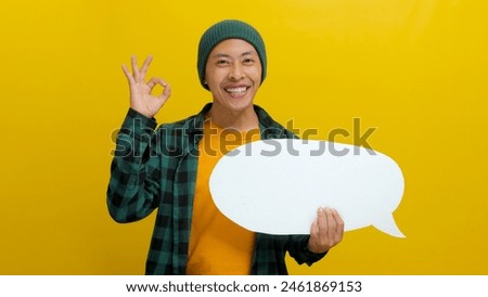 Excited Asian man in a beanie hat and casual shirt shows an OK gesture to the camera while holding a speech bubble with copy space. Isolated on a yellow background