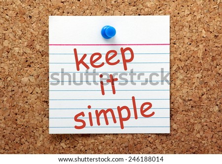 The phrase Keep It Simple in red ink on a white index card pinned to a cork notice board as a reminder