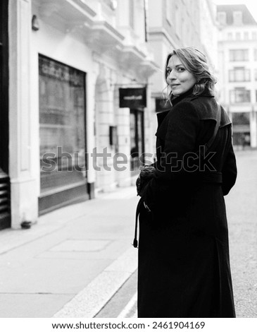 Monochrome classic portrait of woman walking in a London street. She is wearing a black coat and she is showing her back and looking at camera. Film and analogue photography. Grain.